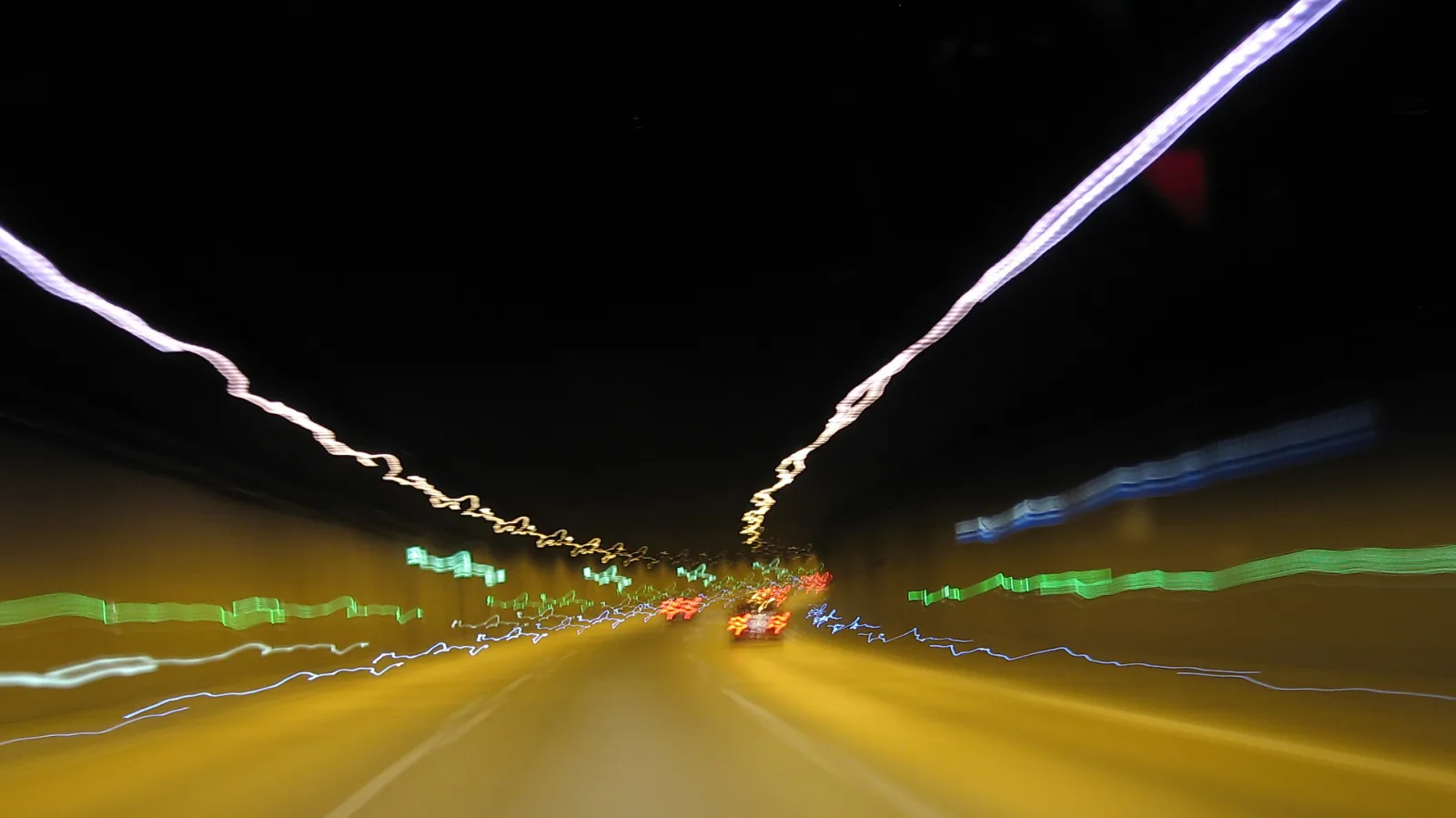 Nächtliche Autobahn mit bunten Lichtstreifen in Grün, Blau und Weiß, in der Ferne rote Rücklichter von Autos, darüber schwarzer Himmel.