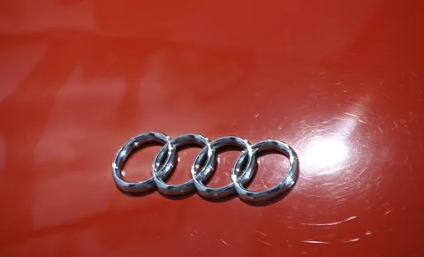 Silberne Audi-Ringe, vier ineinander verschlungene Kreise, auf einer glänzenden roten Karosserie mit feinen Kratzspuren und Lichtreflex.