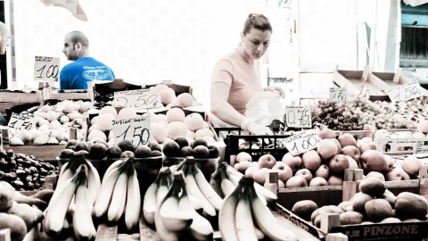 Obststand auf einem Wochenmarkt mit Bananen, Äpfeln und Trauben, eine Verkäuferin in rosa Shirt packt eine Tüte, im Hintergrund ein Mann in blauem Shirt.