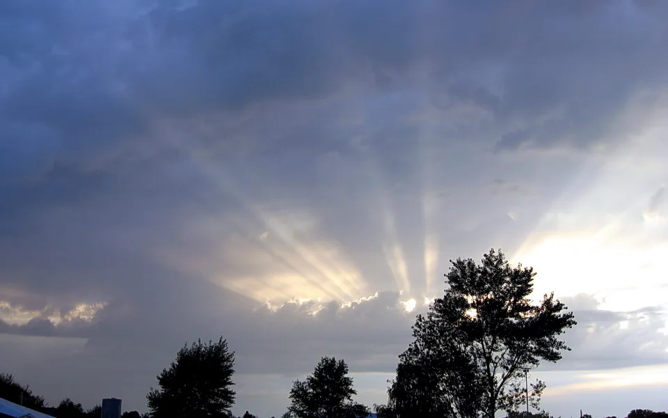 Dunkle Wolkendecke am Abend, Sonnenstrahlen brechen durch die Wolken, im Vordergrund Silhouetten von Bäumen und Zeltdächern.