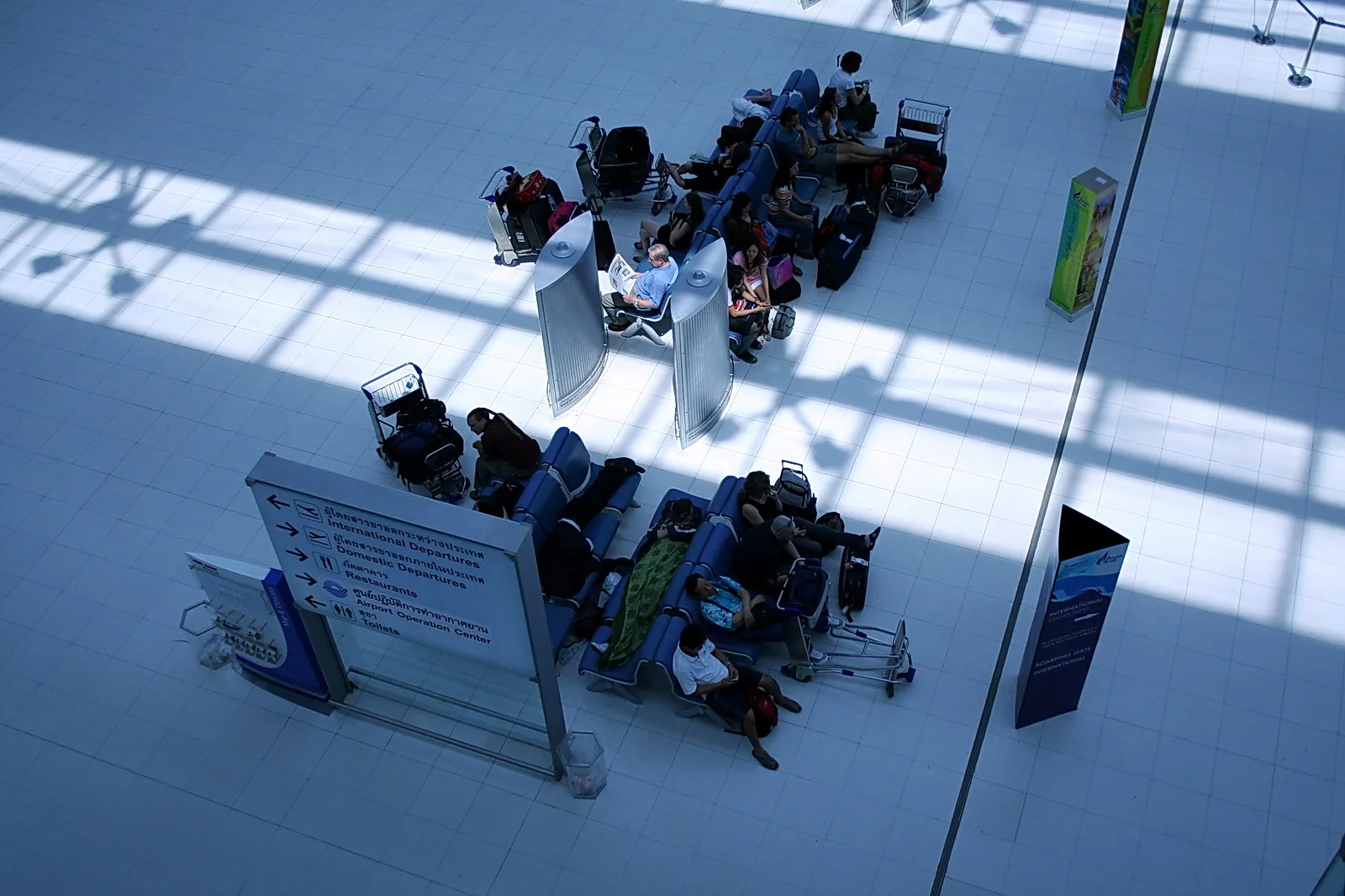 Reisende sitzen auf blauen Bänken in einer hellen Wartehalle, daneben stehen Gepäckwagen, und Schatten von Fensterstreben liegen auf dem Boden.
