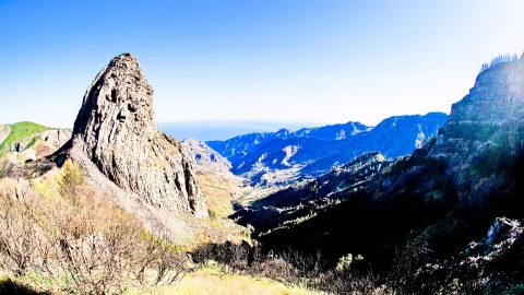 Ein spitzer, heller Felsturm links im Bild vor einem tief eingeschnittenen Bergtal unter wolkenlosem blauem Himmel, im Vordergrund trockenes Gestrüpp.
