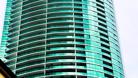 Fassade eines runden Hochhauses mit geschwungenen Balkonreihen und türkisfarbenen Glaselementen vor hellem Himmel
