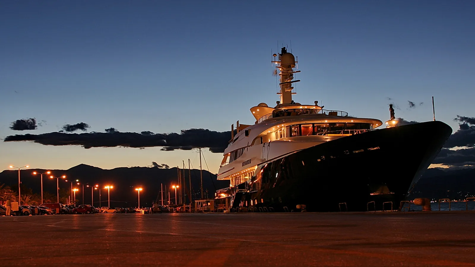 Große weiße Luxusyacht mit beleuchteten Decks liegt abends an der Kaimauer, links Straßenlaternen und parkende Autos, dahinter Berge und dunkler Himmel.