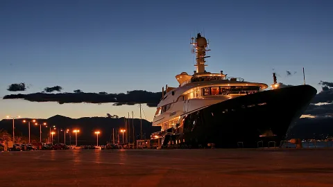 Große weiße Luxusyacht mit beleuchteten Decks liegt abends an der Kaimauer, links Straßenlaternen und parkende Autos, dahinter Berge und dunkler Himmel.
