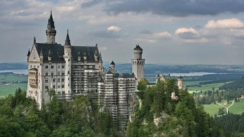 Schloss Neuschwanstein auf einem bewaldeten Felsen, teils von einem Baugerüst umgeben, mit Wiesen und einem See im Hintergrund unter bewölktem Himmel.