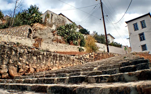 Breite Steintreppe steigt diagonal ins Bild hinauf, links eine Natursteinmauer, rechts weiß getünchte Häuser, oben Bäume und Kaktus vor bewölktem Himmel, ein Mensch steigt auf der Treppe hinauf.