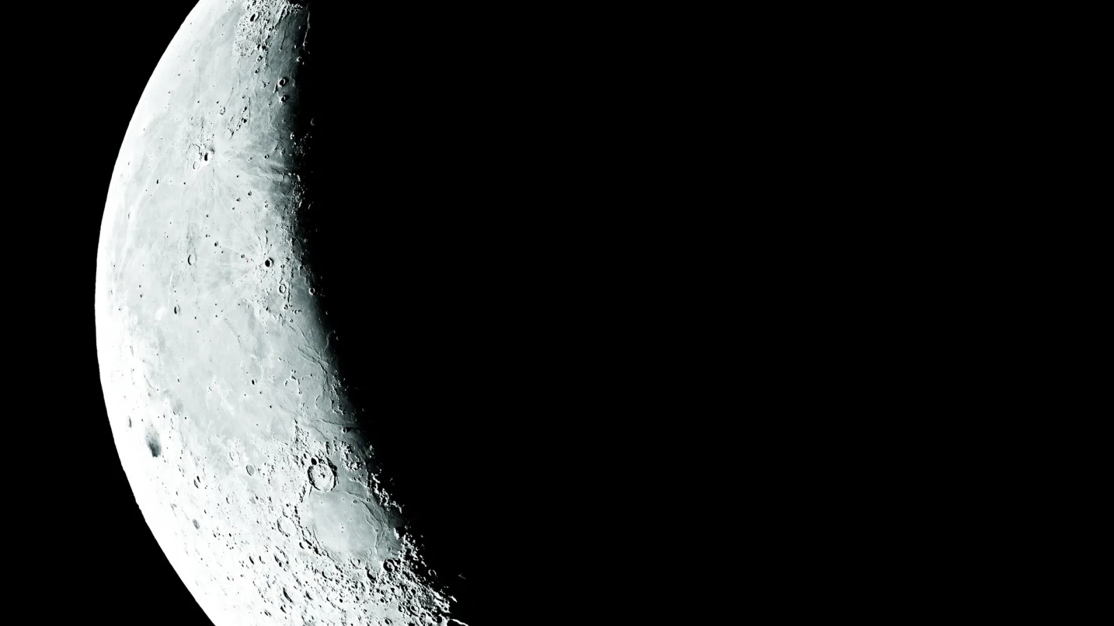 Eine Mondsichel mit zahlreichen Kratern leuchtet links im Bild, der rechte Teil liegt im schwarzen Weltraum.