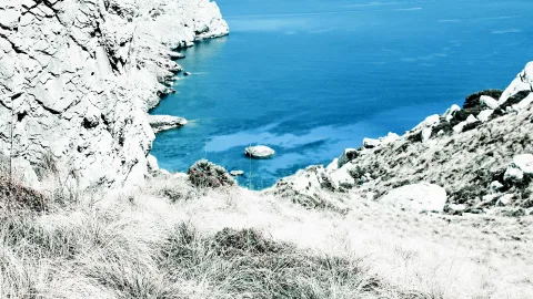 Felsige Küste mit weiß verfärbten Klippen, im Vordergrund trockenes Gras, rechts Felsbrocken, in der Bucht ein kleiner Felsen im tiefblauen Meer.