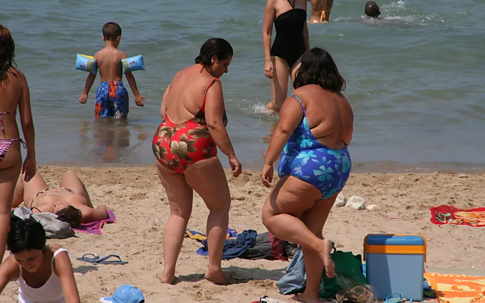 Sommerlicher Strand mit mehreren Badegästen: Zwei Frauen in bunten Badeanzügen stehen im Sand, ein Junge mit Schwimmflügeln läuft im flachen Wasser, im Hintergrund schwimmen weitere Personen.
