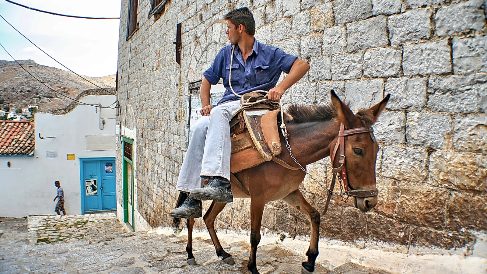Ein Mann in blauem Hemd reitet auf einem braunen Esel eine gepflasterte Steintreppe hinauf, links steht ein weiß getünchtes Haus mit blauer Tür.