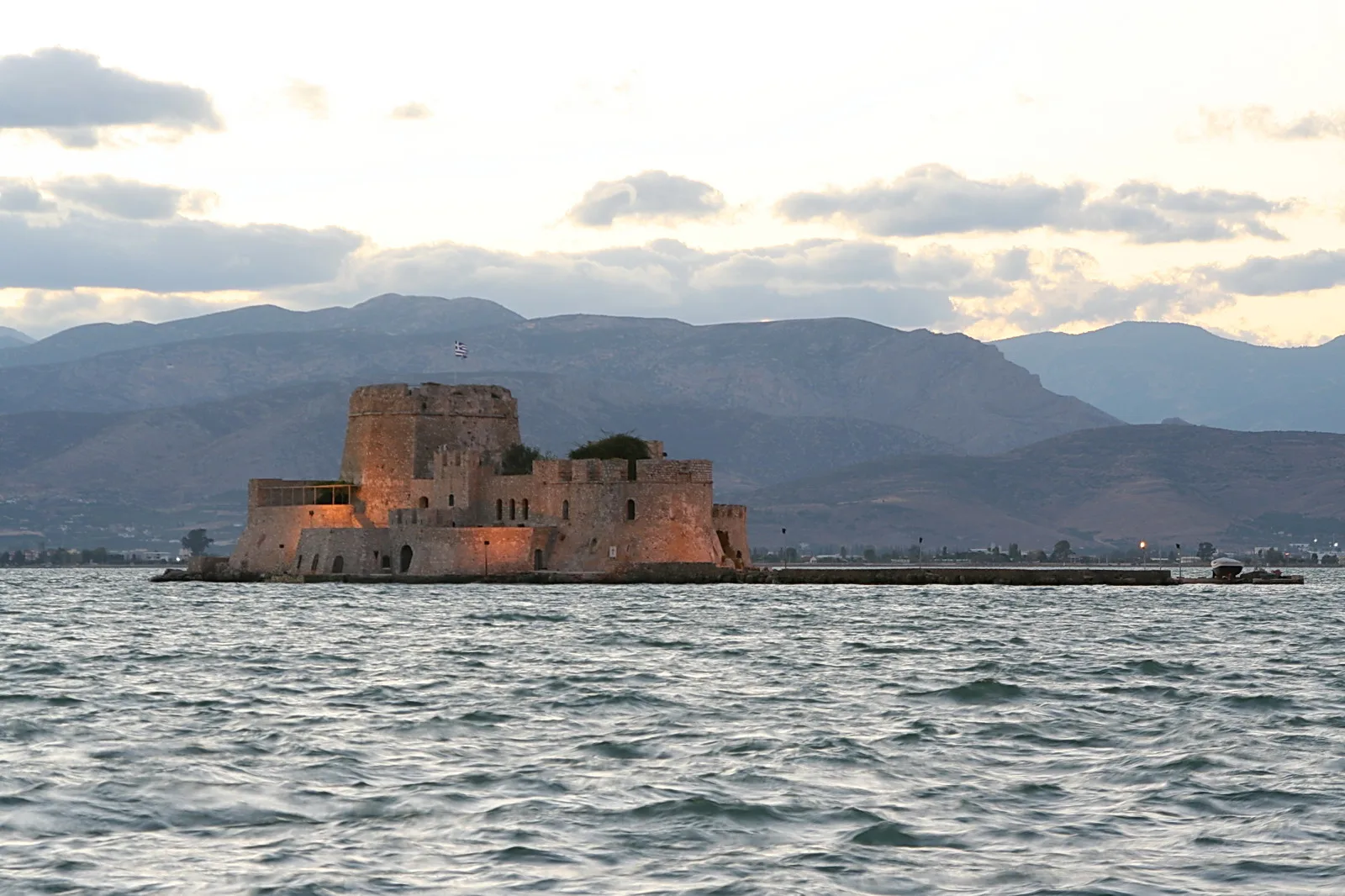 Beleuchtete Festung mit runden Türmen auf einer Insel im Wasser, dahinter Berge unter bewölktem Abendhimmel