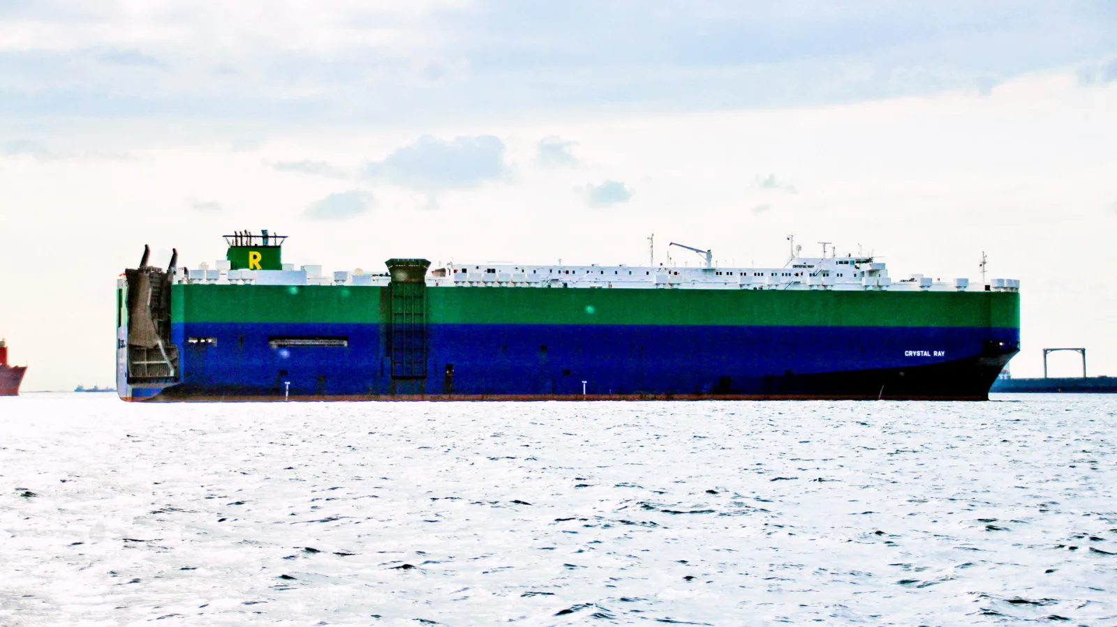 Großer Autotransporter mit grün-blauem Rumpf und weißen Aufbauten liegt ruhig auf dem Wasser, darunter bewölkter Himmel.