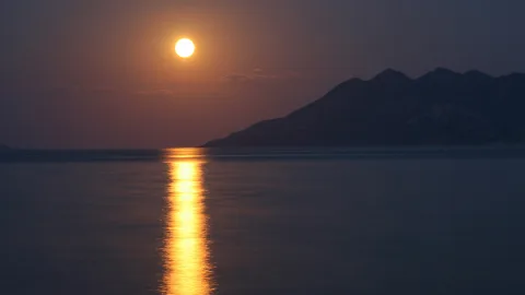 Heller Vollmond über einem ruhigen Meer, rechts dunkle Berge, eine goldene Lichtspur auf dem Wasser.