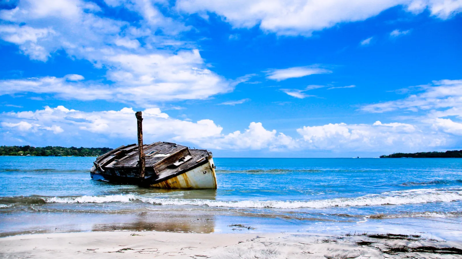 Ein verrostetes, halb verfallenes Holzboot mit weiß-gelbem Rumpf liegt auf flachem Sandstrand, im türkisfarbenen Meer stehen kleine Wellen, darüber ein blauer Himmel mit weißen Wolken.