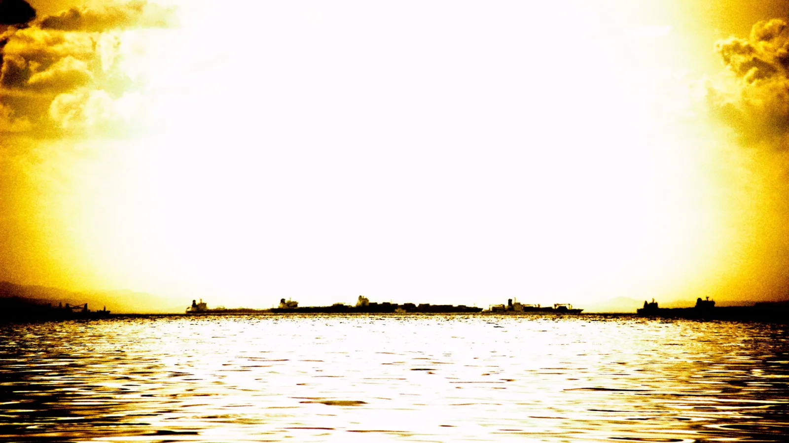 Silhouetten von Frachtschiffen am Horizont über einer goldgelb leuchtenden Wasserfläche, darüber überstrahlter Himmel mit Wolken.