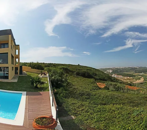 Gelbe zweistöckige Villa mit großen Fenstern und einem blauen Pool auf einer Holzterrasse, dahinter ein grüner Hang und Blick auf eine Küstenlandschaft mit weißen Häusern unter blauem Himmel mit Wolken.
