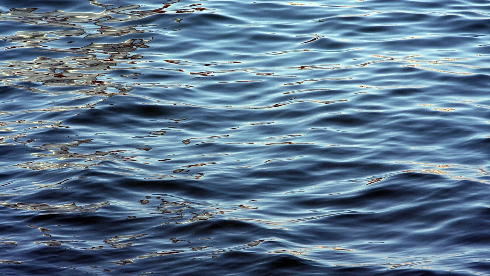 Nahaufnahme einer bewegten Wasseroberfläche mit dunkelblauen Wellen und goldfarbenen Lichtreflexen, besonders links oben.