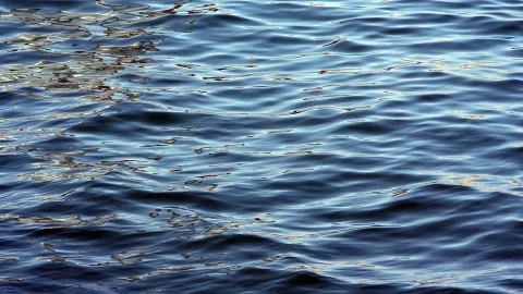 Nahaufnahme einer bewegten Wasseroberfläche mit dunkelblauen Wellen und goldfarbenen Lichtreflexen, besonders links oben.