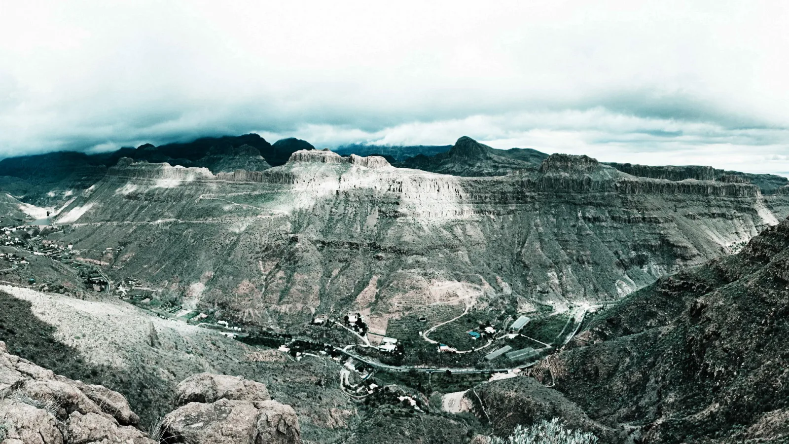 Panoramablick auf eine tief eingeschnittene Bergschlucht mit steilen, terrassierten Felswänden, kleinen Dörfern im Talgrund und dunklen Wolken am Himmel.