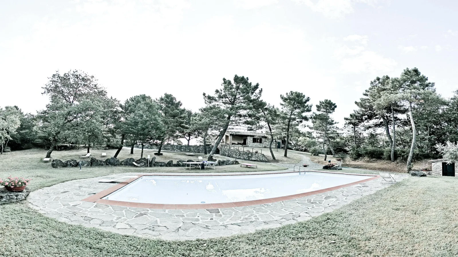 Ovales Swimmingpool mit rotbraunem Rand und Natursteinplatten, dahinter Kiefern, Wiese und ein steinernes Haus unter hellem Himmel.