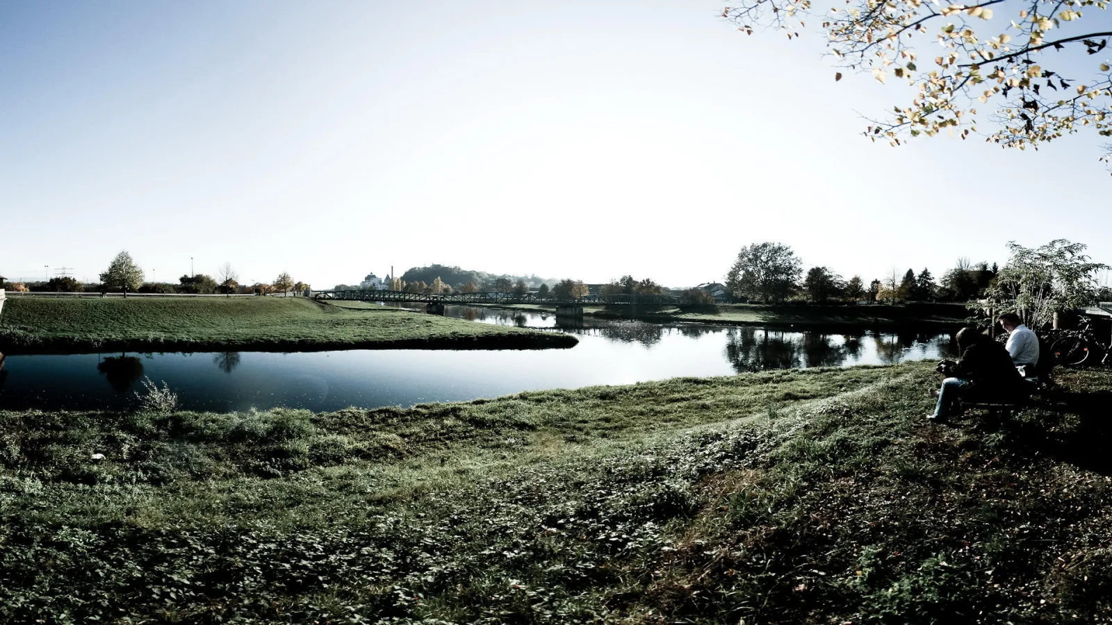 Breiter Fluss unter klarem Himmel mit grüner Uferwiese im Vordergrund, zwei Personen auf einer Bank am rechten Rand, im Hintergrund eine Brücke und Bäume mit herbstlichem Laub.
