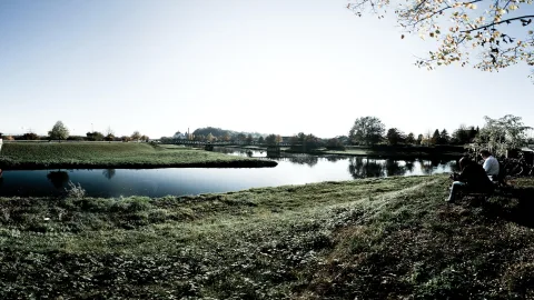 Breiter Fluss unter klarem Himmel mit grüner Uferwiese im Vordergrund, zwei Personen auf einer Bank am rechten Rand, im Hintergrund eine Brücke und Bäume mit herbstlichem Laub.