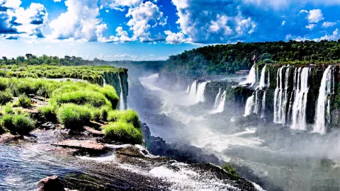 Breites Panorama der Iguazú-Wasserfälle mit weißer Gischt, Nebel über der Schlucht, grüner Vegetation im Vordergrund und blauem Himmel mit Wolken.