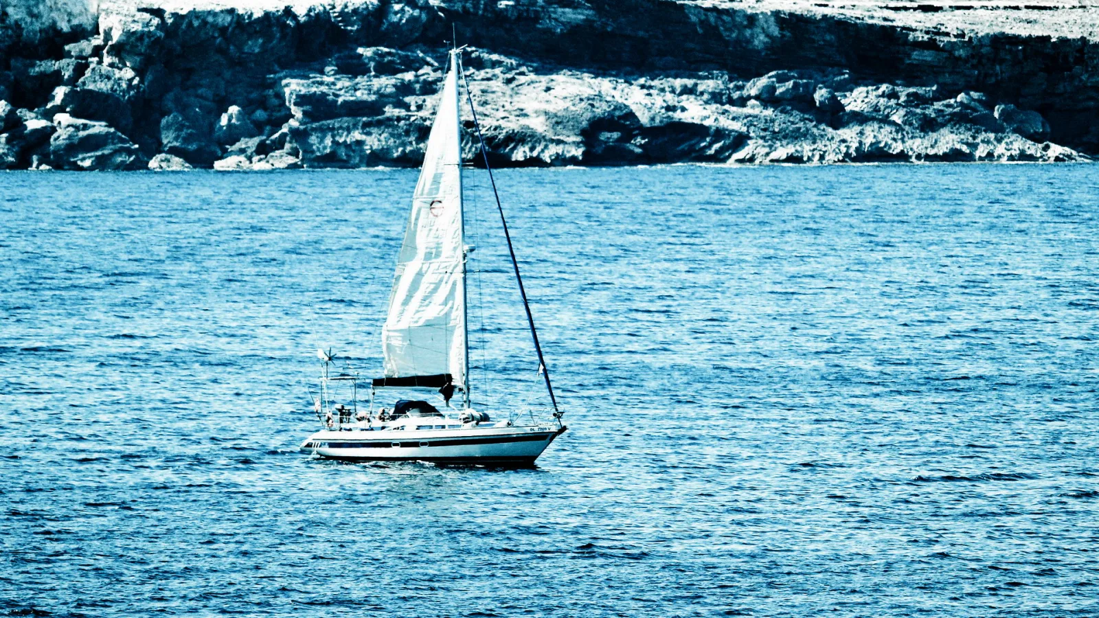 Ein weißes Segelboot mit gesetztem Großsegel fährt auf ruhigem blauem Meer vor einer felsigen Küste.