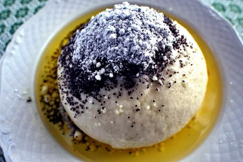 Ein runder Germknödel auf einem weißen Teller, oben mit Mohn und Puderzucker bestreut, umgeben von goldener Vanillesauce.