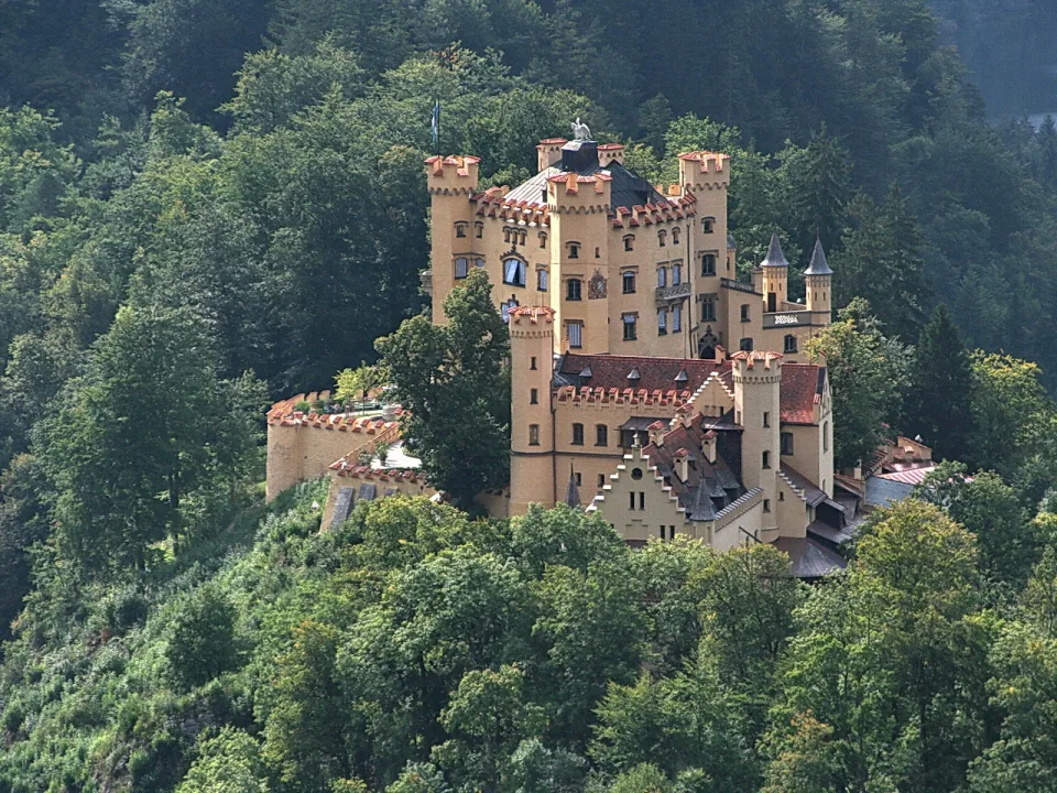 Gelbes Schloss Hohenschwangau mit Zinnen und spitzen Türmen auf einem bewaldeten Hügel, umgeben von dichten Bäumen.