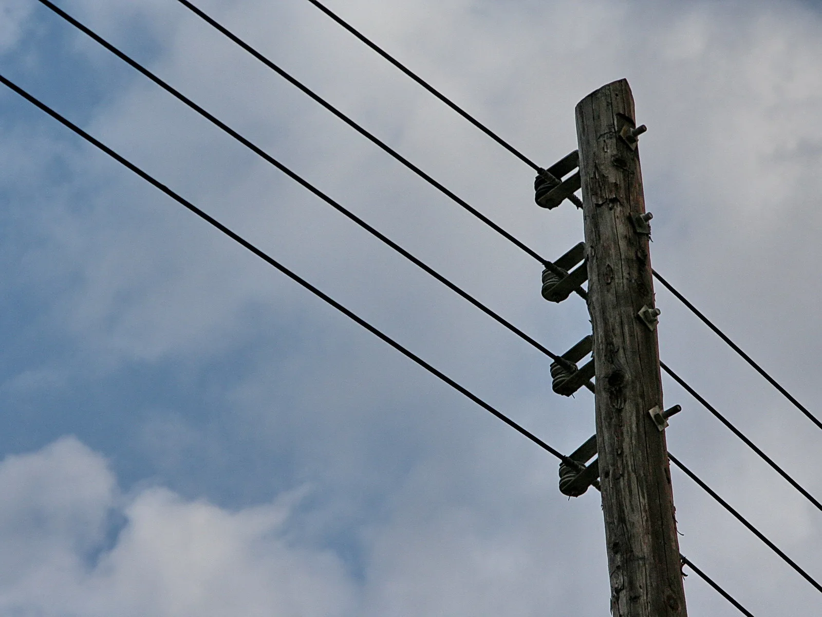 Ein grauer Holzmast ragt diagonal ins Bild, an ihm hängen mehrere schwarze Stromleitungen, die schräg nach links verlaufen. Dahinter ist ein blau-grauer Himmel mit Wolken zu sehen.