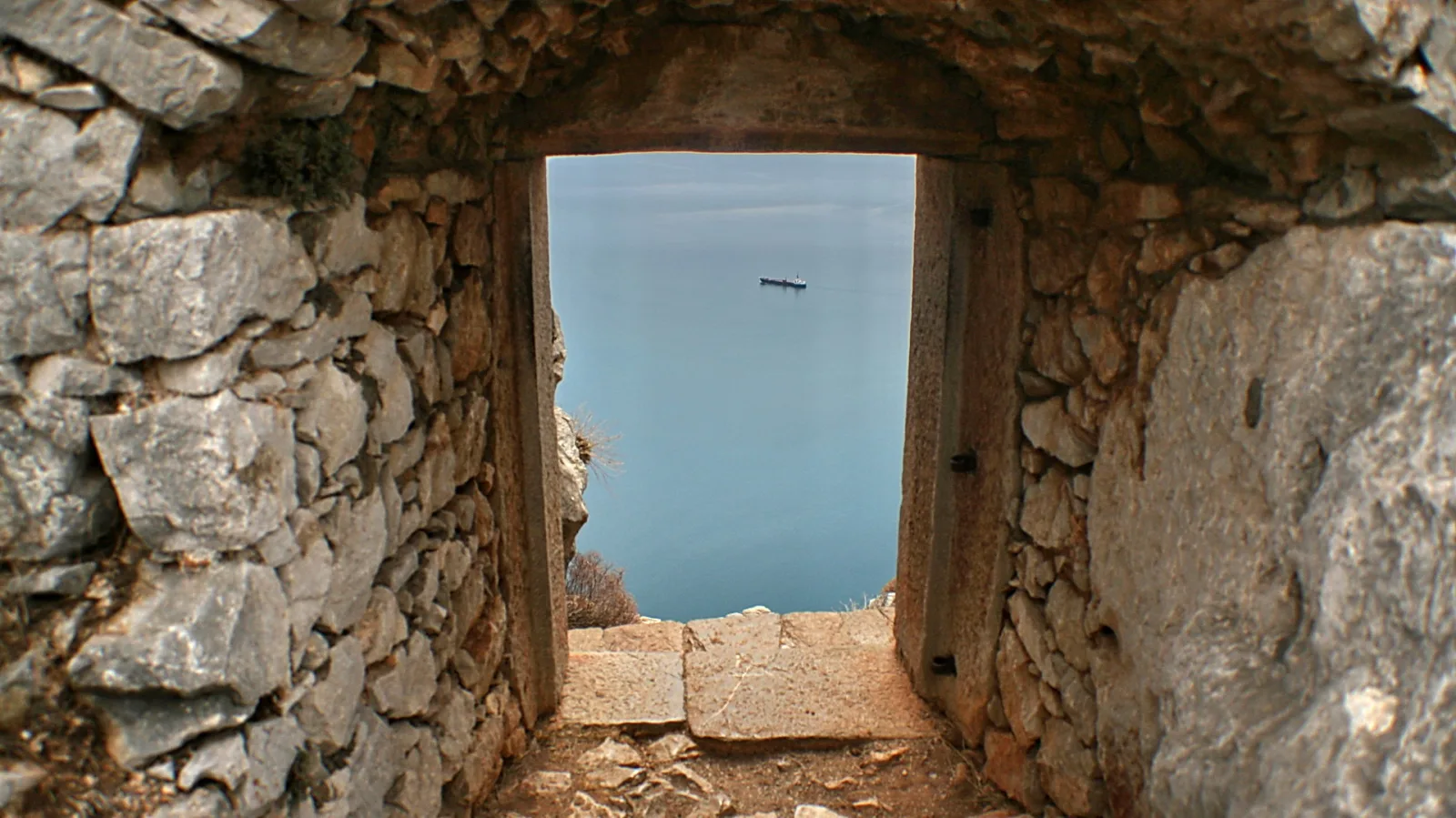 Durchgang in einer alten Bruchsteinmauer, durch den der Blick auf ruhiges blaugraues Meer fällt; in der Ferne ist ein kleines Boot am Horizont zu sehen.