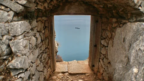 Durchgang in einer alten Bruchsteinmauer, durch den der Blick auf ruhiges blaugraues Meer fällt; in der Ferne ist ein kleines Boot am Horizont zu sehen.