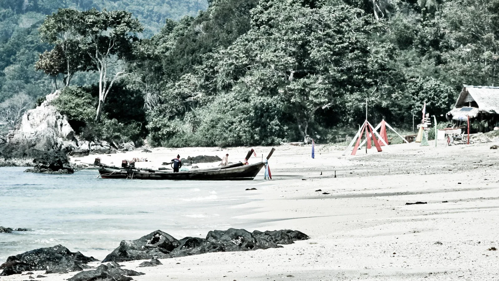 Ein langes Holzboot liegt am Sandstrand, dahinter dichter Wald und eine einfache Strandhütte mit Strohdach.