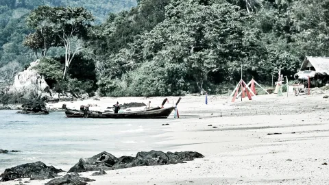 Ein langes Holzboot liegt am Sandstrand, dahinter dichter Wald und eine einfache Strandhütte mit Strohdach.