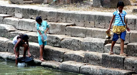 Drei Kinder stehen und hocken auf breiten Steinstufen am Wasser. Ein Mädchen links hält ein Netz ins Wasser, ein Kind in der Mitte trägt eine Plastikflasche, rechts geht ein Junge mit einem Korb die Stufen hinunter.