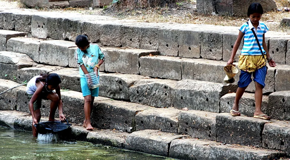 Drei Kinder stehen und hocken auf breiten Steinstufen am Wasser. Ein Mädchen links hält ein Netz ins Wasser, ein Kind in der Mitte trägt eine Plastikflasche, rechts geht ein Junge mit einem Korb die Stufen hinunter.