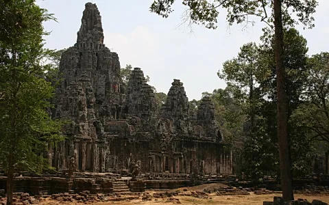 Verwitterter Steintempel des Bayon in Angkor Thom mit mehreren gestuften Türmen, davor Bäume und Steinreste am Boden.