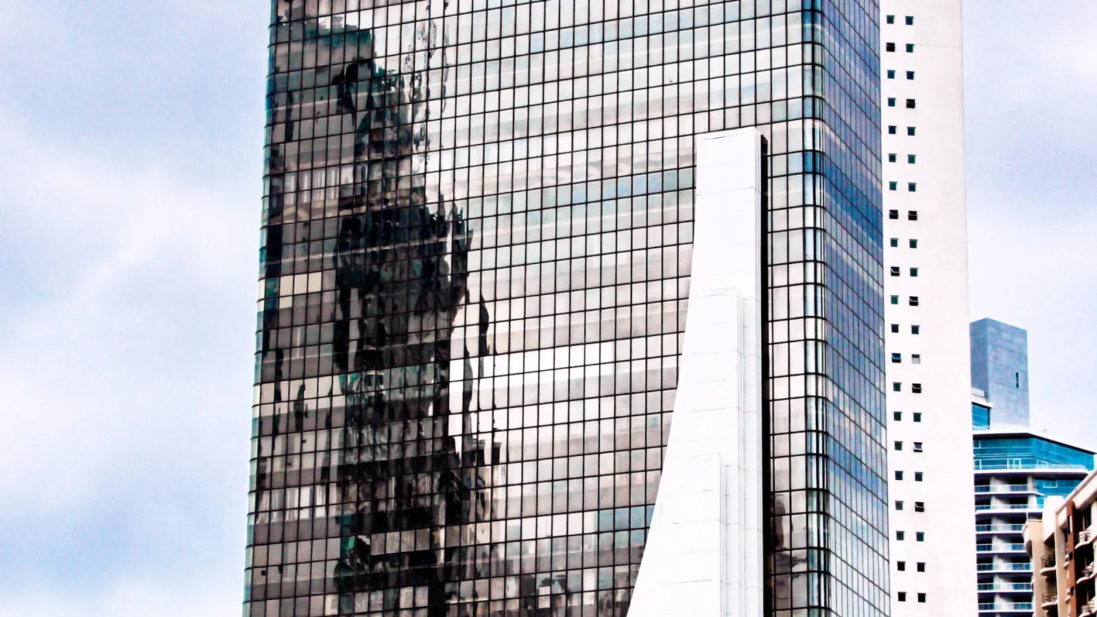 Spiegelnde Glasfassade eines dunklen Hochhauses mit Fensterraster, daneben ein helles weißes Gebäude und blasser Himmel mit Wolken.