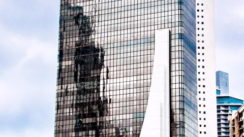 Spiegelnde Glasfassade eines dunklen Hochhauses mit Fensterraster, daneben ein helles weißes Gebäude und blasser Himmel mit Wolken.