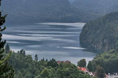 Ein langgestreckter Bergsee zwischen bewaldeten Hängen, im Vordergrund Nadelbäume und einige rote Dächer am Ufer, die Wasseroberfläche spiegelt das diffuse Tageslicht.