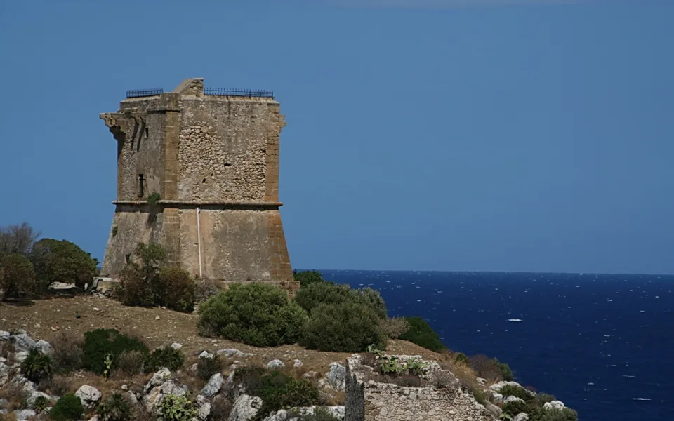 Ein verwitterter Steinturm auf einer felsigen Küste mit Büschen im Vordergrund, dahinter tiefblaues Meer unter klarem Himmel.