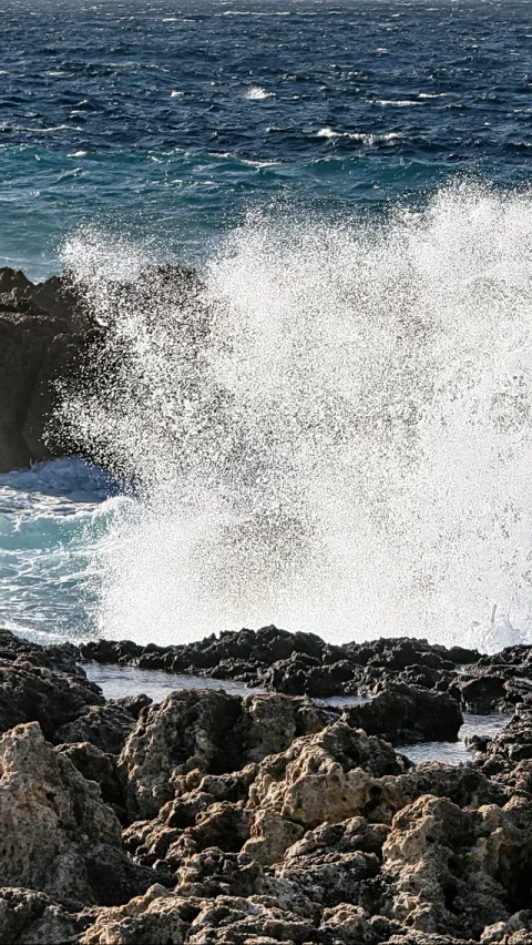 Eine große weiße Welle bricht über dunklen, zerklüfteten Felsen, Gischt spritzt in die Luft, dahinter liegt dunkelblaues Meer.