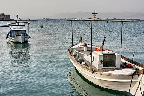 Zwei weiße Boote liegen auf ruhigem, blaugrauem Wasser, das Boot rechts mit Holzdeck und Metallgestänge, im Hintergrund Dunst, Berge und eine Festungsmauer.