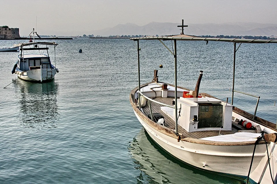 Zwei weiße Boote liegen auf ruhigem, blaugrauem Wasser, das Boot rechts mit Holzdeck und Metallgestänge, im Hintergrund Dunst, Berge und eine Festungsmauer.