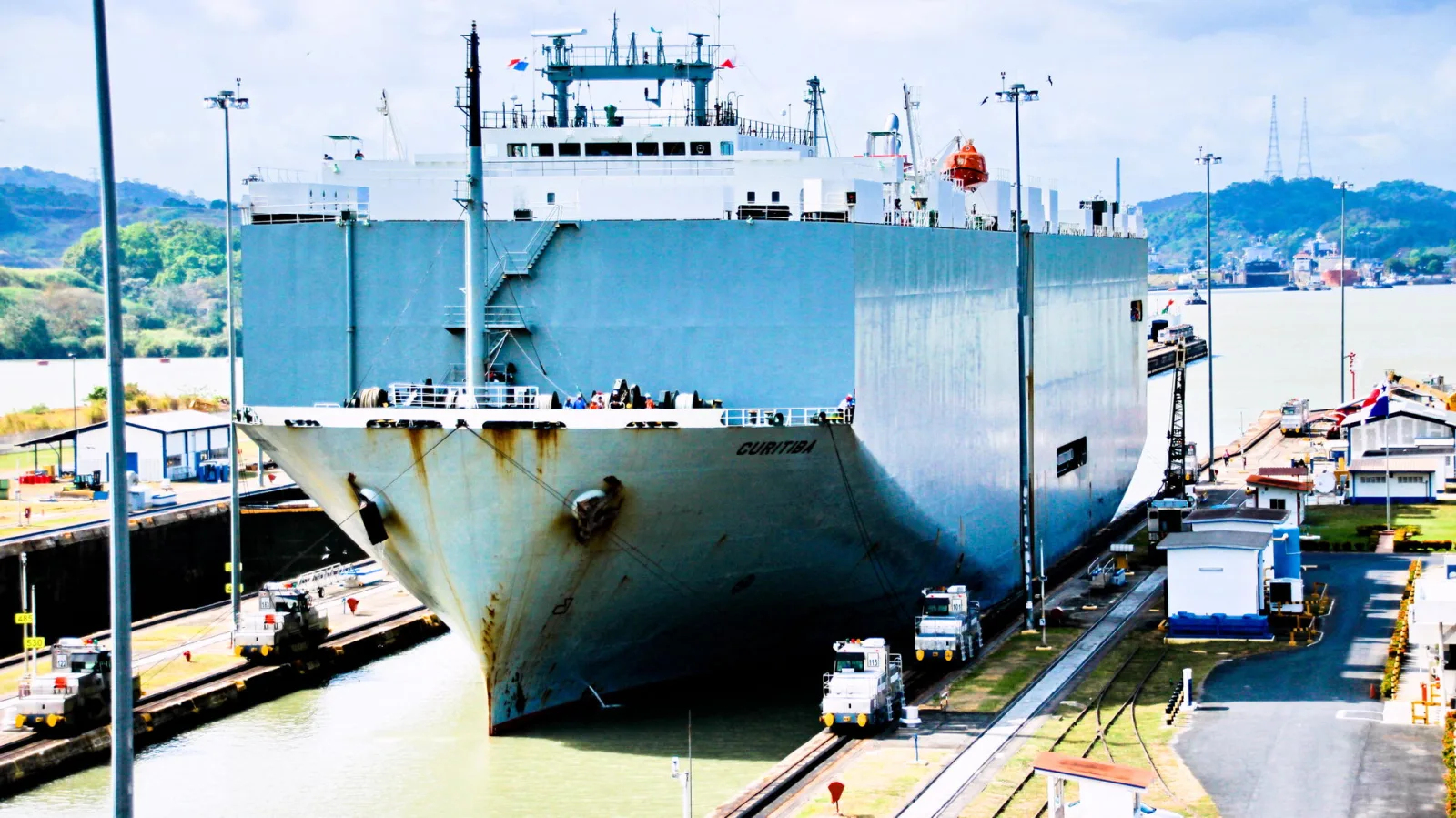 Großer grauer Frachter Curitiba fährt in eine Schleuse des Panamakanals, links und rechts ziehen Schlepploks das Schiff.