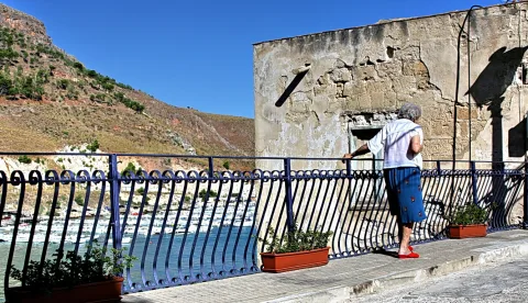 Ältere Frau in weißer Bluse und blauem Rock steht an einem blauen schmiedeeisernen Geländer und blickt auf einen Hafen mit Booten. Links ein steiler, bewachsener Hang, rechts eine verwitterte Steinmauer unter blauem Himmel.