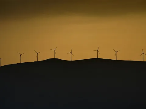 Silhouetten mehrerer Windräder auf einem dunklen Hügelkamm vor orangefarbenem Abendhimmel