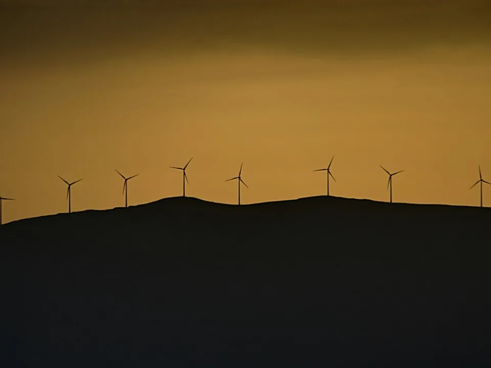Silhouetten mehrerer Windräder auf einem dunklen Hügelkamm vor orangefarbenem Abendhimmel